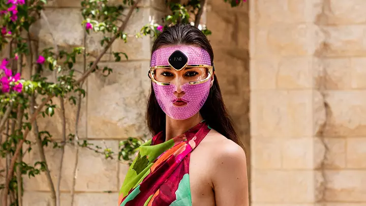 A gorgeous woman with dark hair and dark eyes, in a summer dress, in front of purple flowers, wearing a led face mask set on a purple light 